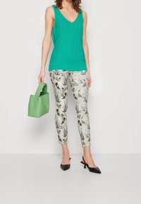 Femme en top tricot sans manches teal, pantalon slim à motif floral, escarpins noirs à bride arrière, tenant un petit sac à main vert.