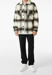 Rutigt flanelljacka i krämfärg och mörkbrunt med två bröstfickor, knäppning fram och en avslappnad passform, i kombination med svarta byxor och vita sneakers.