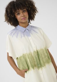 Kreminės spalvos trumpomis rankovėmis marškiniai su sagomis, kuriuose šviesiai mėlynas apykaklė ir žalia tinklinė spalva, su pleištinėmis kišenėmis ir laisvu siluetu.