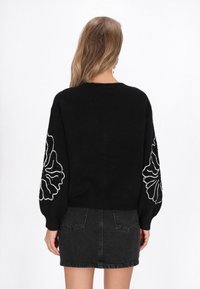 Fekete kötött pulóver, amelyet puffos ujjak díszítenek, mindkét karon fehér virágmintával. Sötét farmer szoknyával viselve.