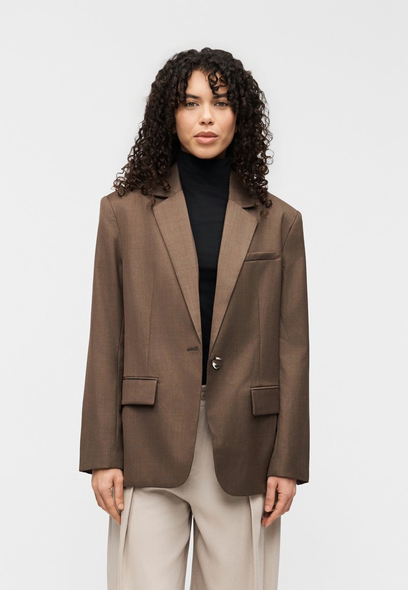 Καφέ σακάκι ραμμένο στα μέτρα με γιακά με εγκοπή, κούμπωμα με ένα κουμπί και δύο μπροστινές τσέπες. Φορεμένο πάνω από μαύρο ζιβάγκο. Ομαλή υφή.