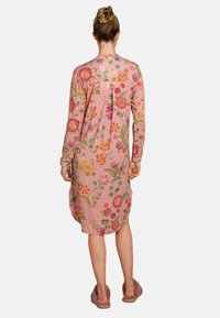 Vrouw met haar in een bun, gekleed in een lange roze bloemenjurk tot de knie en mauve slippers, staande met haar rug naar de camera tegen een witte achtergrond.