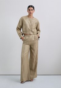 Chaqueta corta beige con dobladillo elástico, combinada con pantalones de pierna ancha, ambos con patrones de hilo metálico y una textura suave.