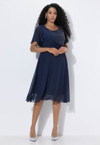 Navyblaues Kleid mit transparenten kurzen Ärmeln, einem strukturierten Oberteil und einem fließenden Rock, kombiniert mit silbernen High Heels.