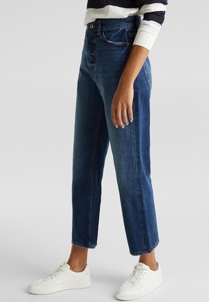 Personne portant un jean en denim bleu taille haute et des baskets blanches, debout devant un fond gris clair uni.