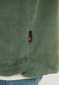 Vihreä flanellitakki teksturoidulla pinnalla, jossa on musta etiketti oranssilla korostuksella helman läheisyydessä. Näkyvät ompeluvastat.