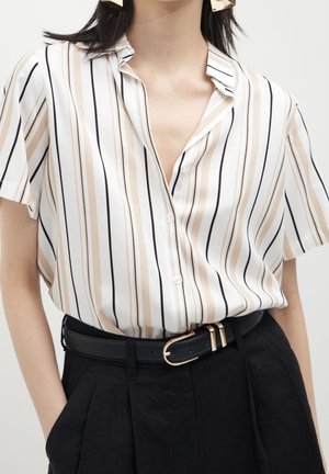 Kvinde iført en kortærmet, vertikalt striped bluse, der er stukket ned i højtaljede sorte bukser med sort bælte og guldbæltespænde.