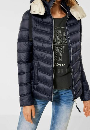 Laivastonsininen untuvatakki, jossa on beige kaulus, vaakasuora ommeltyyli, vetoketjusuljenta ja säädettävät remmit. Päällä tumman t-paidan kanssa.