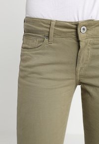 Pantalones de algodón mezclado color caqui con un corte ajustado, diseño de cinco bolsillos, cierre de botón y detalles de costura visibles.