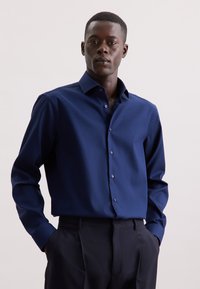 Marineblauw overhemd van gestructureerde stof, met een puntige kraag en een knoopsluiting aan de voorkant, gecombineerd met donkere broek.