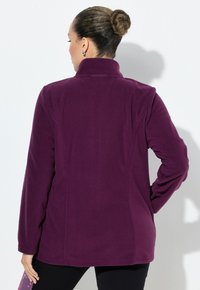 Donna con capelli raccolti in uno chignon, che indossa una giacca in pile viola a maniche lunghe, in piedi con una mano sui fianchi, vista da dietro su uno sfondo bianco.