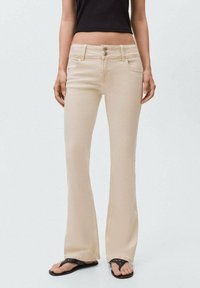 Beige flare jeans iz bombažne mešanice, z zapiranjem na dve gumbni, petimi žepi in mehko teksturo. Nosi se z črnimi natikači.