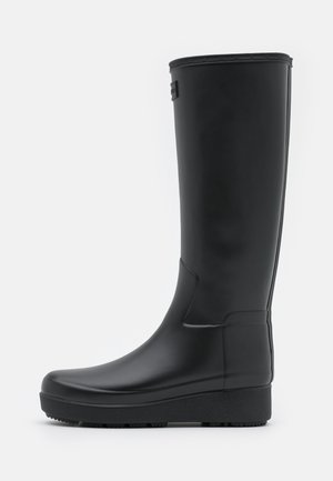 Botte en caoutchouc noire montante jusqu’au genou avec une semelle plate texturée et une surface lisse, vue de profil sur fond blanc.