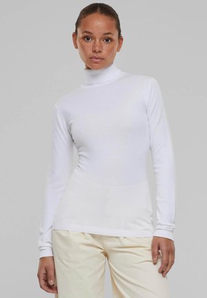 White long-sleeve turtleneck top with a fitted design, ribbed texture at cuffs and hem, and smooth fabric. Paired with light-colored trousers.