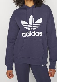 Mörkblå huvtröja med en stor vit Adidas-logotyp, framficka av kangaroo-modell, dragsko i huvan och ribbade mudd- och nederkanter. Mjuk tygstruktur.