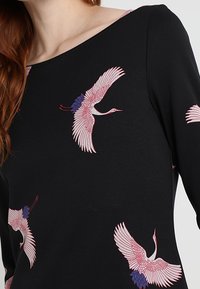 Un haut noir avec un large décolleté, présentant plusieurs grues brodées en rose et bleu dans un motif dynamique de vol sur le tissu.
