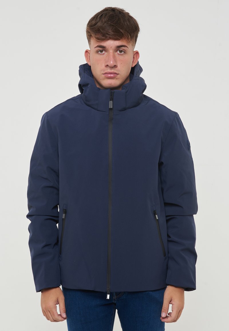 Veste isolante bleu marine avec un col haut, fermeture éclair sur le devant et deux poches latérales. Texture lisse et capuche pour une protection supplémentaire.