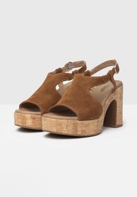 Sandales en daim marron avec un bout ouvert et un talon en liège évasé. Présentent un design à découpes et une sangle de cheville réglable avec une boucle métallique.
