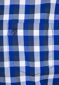 Tissu à carreaux bleu, blanc et gris avec une poche cousue délimitée et un petit logo noir brodé dans la partie supérieure gauche de la poche.