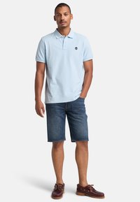 Polo de color azul claro con mangas cortas, combinado con pantalones cortos de mezclilla oscuros. Zapatos de barco marrones con cordones. Logo en la camisa. Diseño simple.