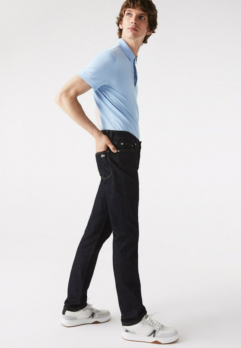 Dunkelblaue Jeans im Slim Fit, kombiniert mit einem hellblauen Poloshirt und weißen Sneakers. Die Jeans besitzen traditionelle Taschen und Nähte.