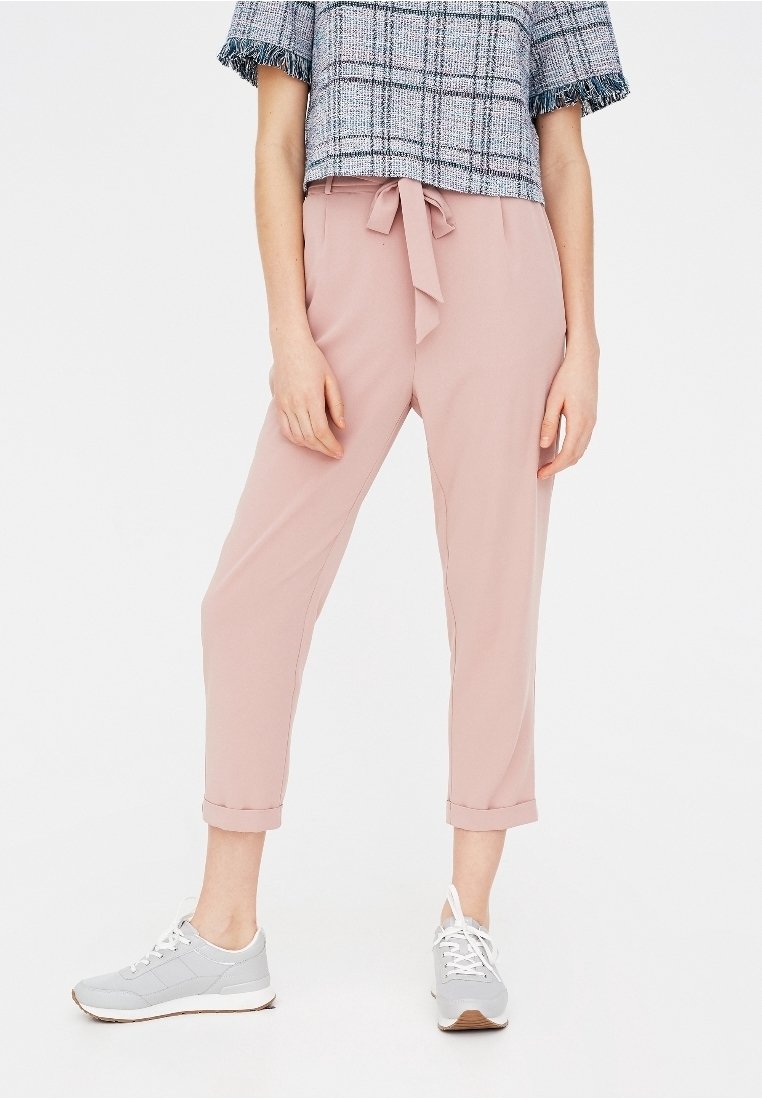 Pantalons tapered rose clair avec un détail de taille nouée, des poignets retroussés et un tissu lisse, associés à des baskets de sport grises.