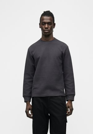 Junger Mann steht, trägt ein dunkelgraues, strukturiertes Langarmshirt und schwarze Hose vor einem schlichten, hellen Hintergrund.