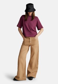 Camicetta corta bordeaux abbinata a pantaloni beige a gamba larga. Il modello indossa un cappello a secchiello nero, con le mani in tasca, mettendo in evidenza il taglio e lo stile degli abiti.