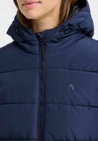 Dunkelblaue Daunenjacke mit hohem Kragen, durchgehendem Reißverschluss und verstellbarem Kordelzug. Besitzt horizontale Nähte und ein Logo auf der Brust.