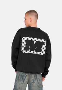 Schwarzes, oversized Sweatshirt mit einem karierten schwarz-weißen Patch, das auf der Rückseite ein großes schwarzes "M" zeigt. Strukturiertes Material und lockere Passform.