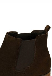 Donkerbruine suède Chelsea boot met elastische zijpanelen en een trekontlasting aan de achterkant, met close-up detail van het bovenste gedeelte.