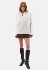 Pull en tricot blanc surdimensionné avec motif en relief, superposé à une chemise blanche, associé à une mini-jupe marron et des bottes noires à talons hauts jusqu'aux genoux.