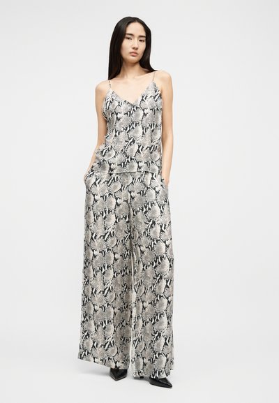 Frau steht mit den Händen in den Taschen und trägt ein passendes graues und schwarzes Top mit Schlangenledermuster sowie eine weit geschnittene Hose, dazu schwarze spitze Schuhe.