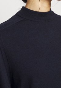 Maglione a collo alto blu navy con texture a coste e design aderente, che mette in mostra le cuciture sulle spalle e una finitura della stoffa liscia.
