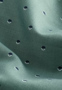 Textil con una tela verde texturizada que presenta pequeños puntos en azul marino y blanco en un patrón. Textura suave y lisa visible.