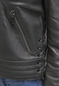 Black leather jacket featuring a zippered pocket, lace-up detailing on the side, and ribbed hemline. Smooth texture with an asymmetrical design.