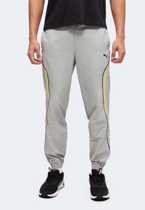 Homme portant un pantalon de jogging Puma gris clair avec des panneaux latéraux jaunes et des baskets noires, debout devant un fond uni.