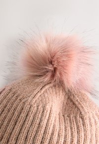 Puha rózsaszín színű kötött sapka bordás textúrával, tetején egy nagy világos rózsaszín szőrmés bojttal, amely kontrasztos részletet ad.