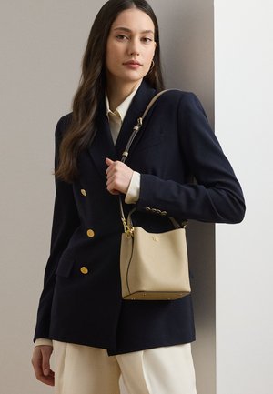 Femeie în sacou bleumarin și pantaloni crem, ținând un geantă tip găleată din piele bej, cu accesorii aurii și curea ajustabilă.