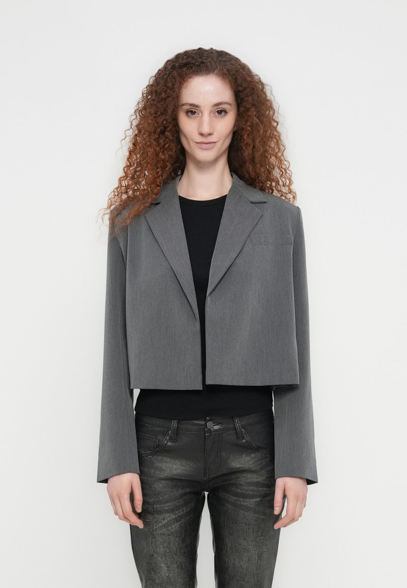 Κοντό γκρι σακάκι με ομαλή υφή, ένα κουμπί και πέτο με εγκοπή, συνδυασμένο με μαύρο τοπ και στενά μαύρα παντελόνια.