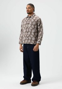 Chemise boutonnée à motifs bruns avec un design floral blanc, associée à un jean large bleu foncé et des chaussures marron à lacets avec des semelles noires.