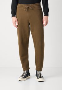 Braune Sweatpants aus weichem Stoff, mit elastischem Bund und Kordelzug, schmal zulaufenden Beinen und Seitentaschen. Getragen mit schwarzen Sneakern.