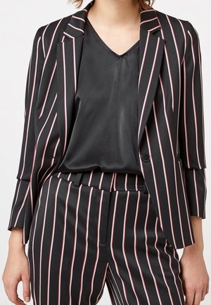 Femme portant un tailleur noir à rayures verticales roses et blanches, associé à un chemisier noir à col en V.