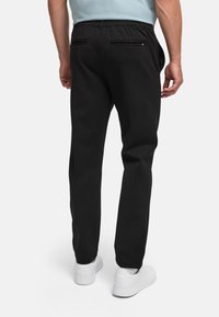 Mann iført svarte bukser og hvite joggesko, står med ryggen til foran en ensfarget bakgrunn.