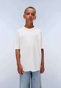 Bílá bavlněná tričko s krátkým rukávem a žebrovaným výstřihem, doplněné s lehkými modrými širokými džíny. Nenápadné logo na hrudi.