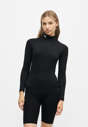 Vrouw draagt een aansluitend zwart topje met lange mouwen en een wit logo op de kraag en zwarte knielange shorts tegen een effen achtergrond.