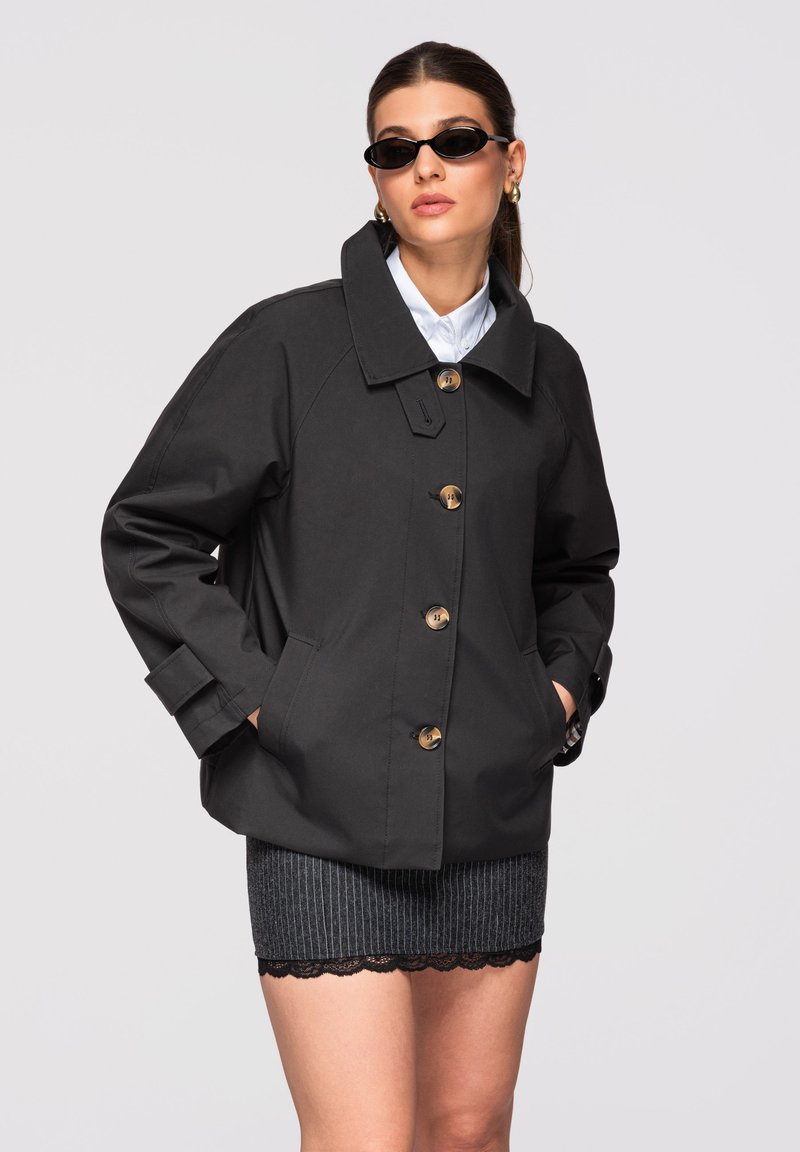 Femme portant une veste noire boutonnée, une chemise blanche à col, une jupe grise à fines rayures avec bordure en dentelle, des lunettes de soleil noires et des boucles d'oreilles en or en forme de créoles.