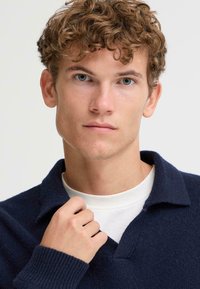 Marineblauwe gebreide trui met een V-hals en kraag; textiel met een zachte uitstraling; model houdt de kraag lichtjes vast met een wit shirt eronder.