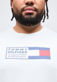 Άνδρας με γενειάδα που φοράει λευκό μπλουζάκι με το λογότυπο και το κείμενο Tommy Hilfiger σε μπλε, λευκό και κόκκινο, που γράφει «All American Classics Est. 1985 New York City».