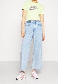 Ljusgrön crop-top med ett leopardmönstrat logo, tillsammans med högmidjade, löst sittande ljusblå denimjeans och vita sneakers med rosa detaljer.